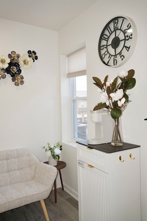 Un salón moderno con un sofá, un reloj decorativo, un jarrón con flores y una ventana con persianas.