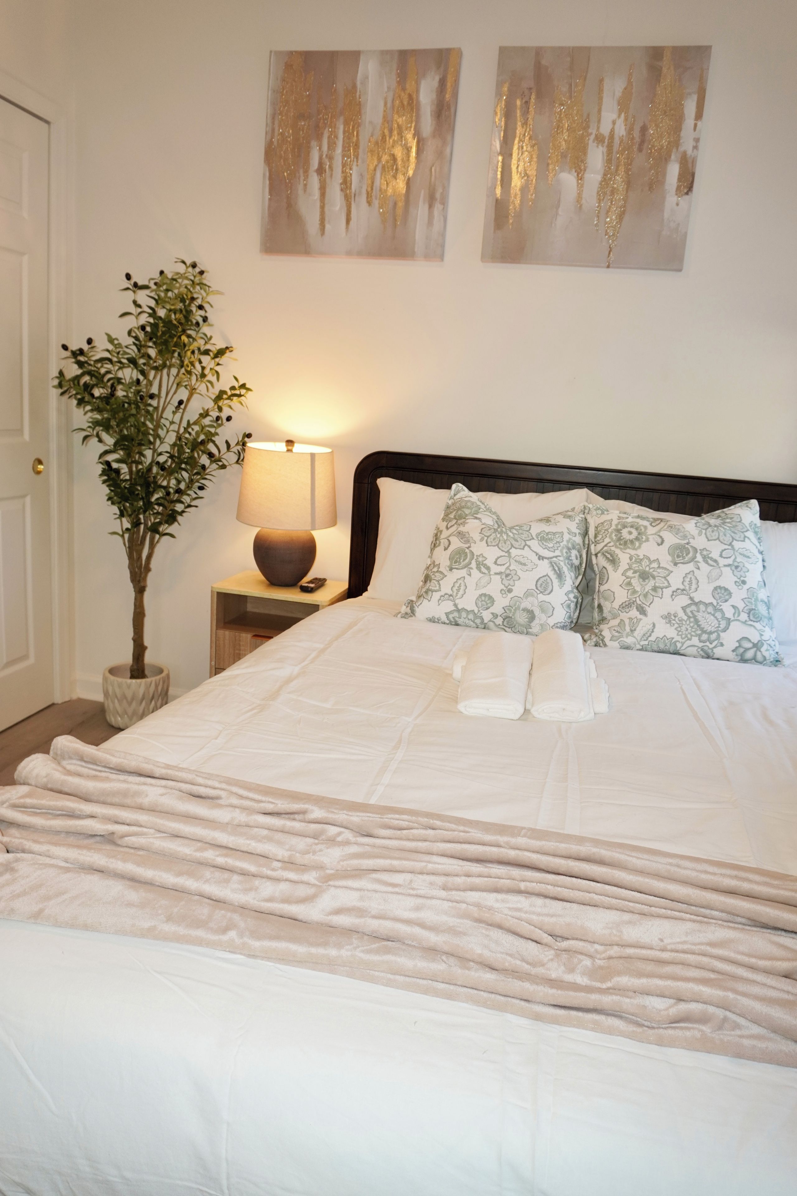 A neatly made bed with a white comforter and two pillows, a bedside lamp, a potted plant, and abstract wall art.