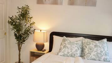 A neatly made bed with a white comforter and two pillows, a bedside lamp, a potted plant, and abstract wall art.