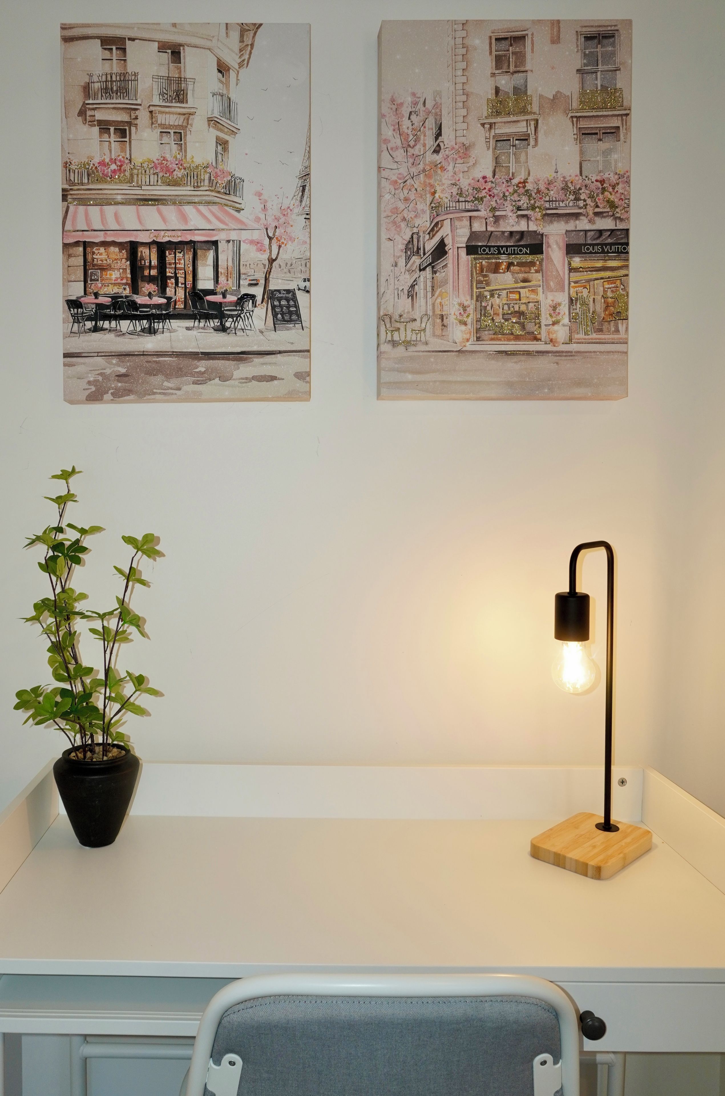 Ett skrivbord med en krukväxt, en lampa och två väggdekorationer som föreställer stadsmotiv.