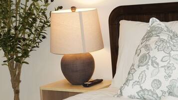 Un lit bien fait, recouvert d’une couette blanche et agrémenté d’un coussin à motifs, une lampe de chevet posée sur une table de nuit en bois, et une plante en pot aux feuilles vertes et aux baies foncées.