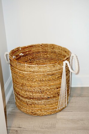 Una cesta tejida con asas, sobre un suelo de madera frente a una pared blanca.