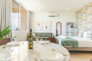 A hotel room with a dining area, a bed with a green bedspread, a mirror, and a wall-mounted air conditioner. - Cica Studio by Atlantic Holiday (Santa Cruz)