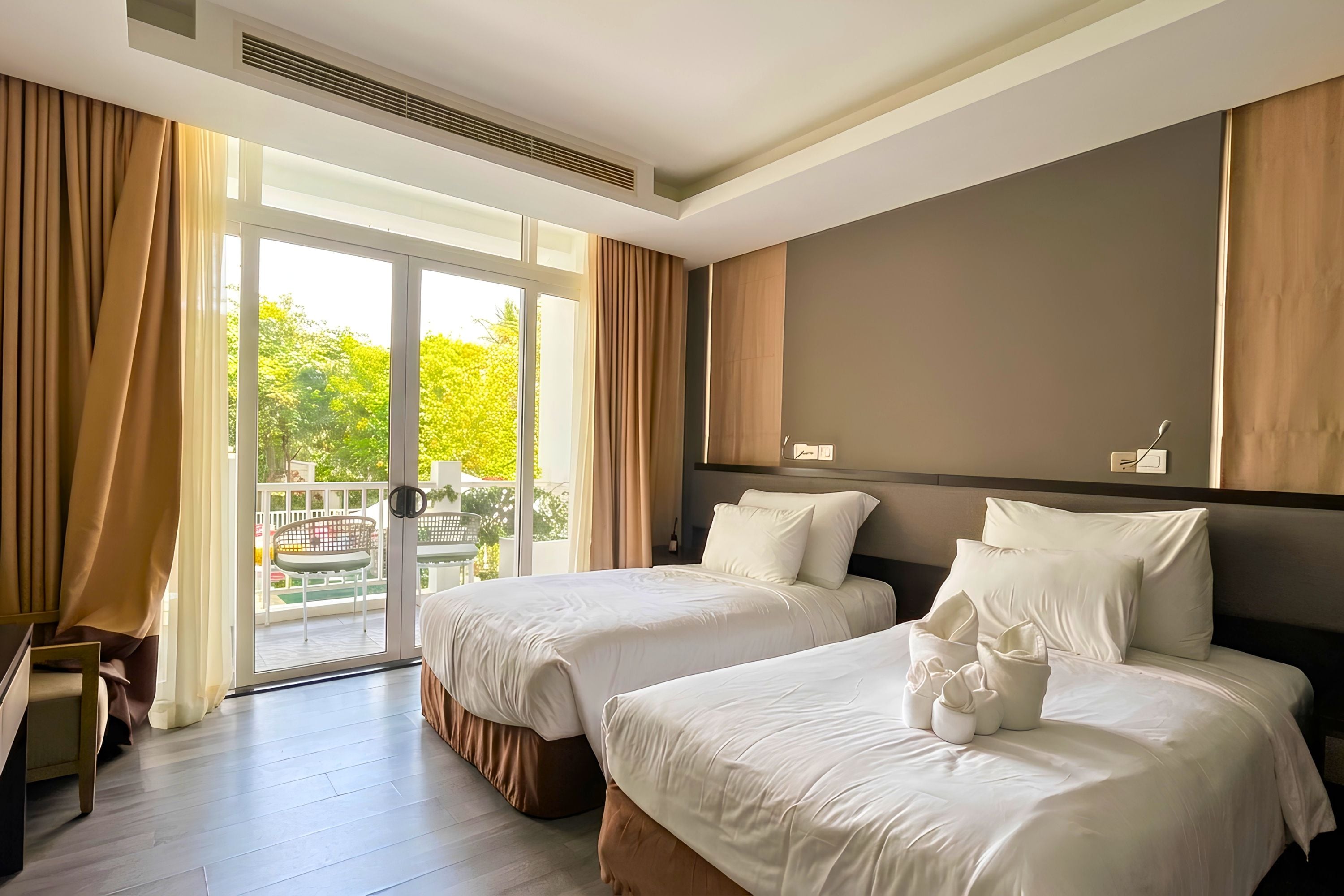 A hotel room with two beds, a large window with curtains, a view of greenery outside, and a ceiling vent.