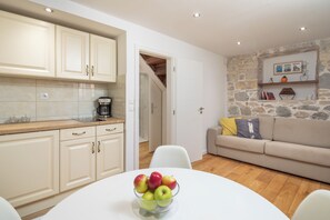 A modern kitchen with white cabinets and a coffee maker, a dining table with a fruit bowl, and a living area with a sofa and stone wall. - C - beautiful modern studio in the heart of Split (Split)