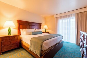 Ein Hotelzimmer mit einem großen Bett, zwei Nachttischen inklusive Lampen, einem Holzkopfende und einem blau gemusterten Teppich. Ein Fenster mit Vorhängen ist ebenfalls zu sehen.