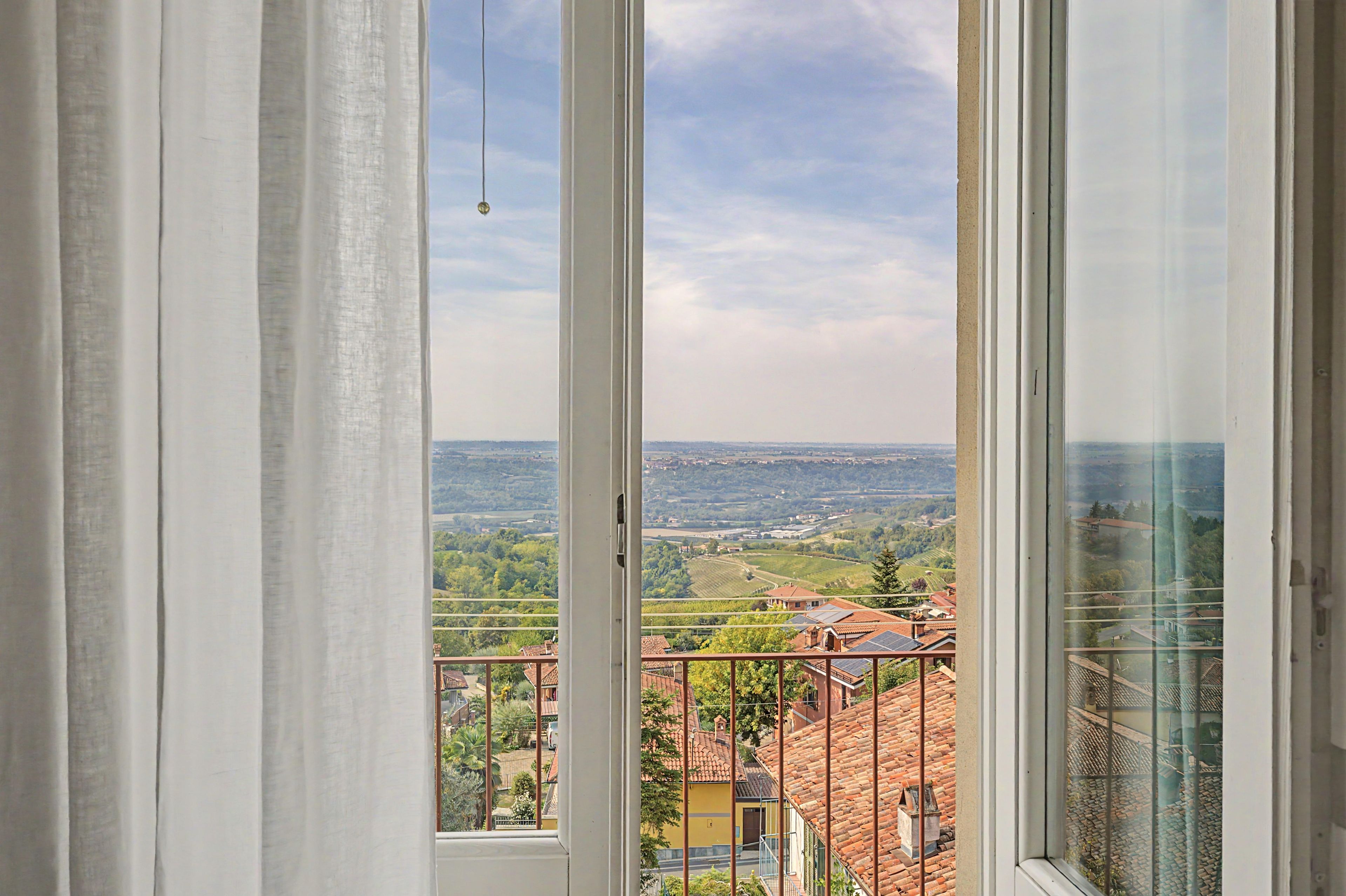Blick aus einem Fenster mit Vorhängen auf eine Landschaft mit Gebäuden und Grünpflanzen.