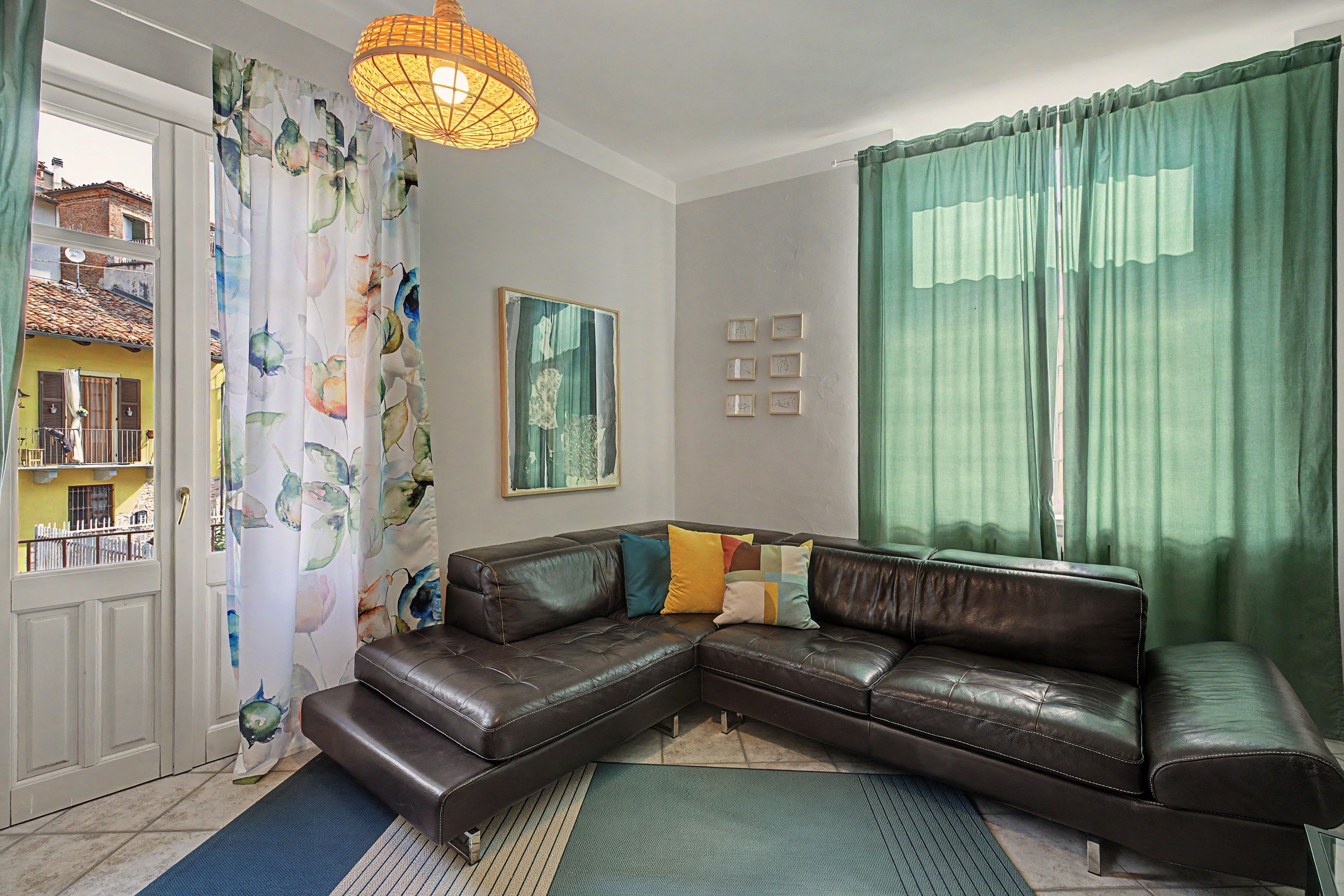 A living room with a brown leather sofa, a patterned curtain, a green curtain, a hanging light fixture, and a painting on the wall.