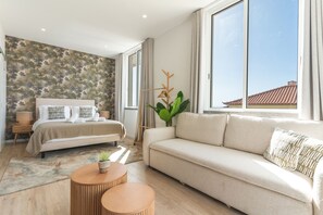 Ein modernes Schlafzimmer mit einem Bett, einem Sofa, einem Nachttisch, einer Lampe, einer Pflanze und einem großen Fenster.
