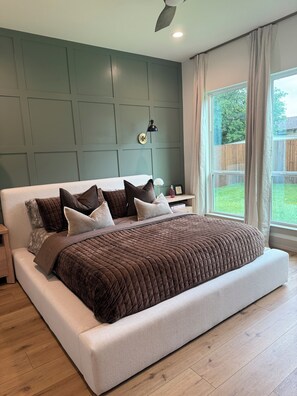 A modern bedroom with a large bed, a nightstand, a wall-mounted lamp, and a window with curtains.