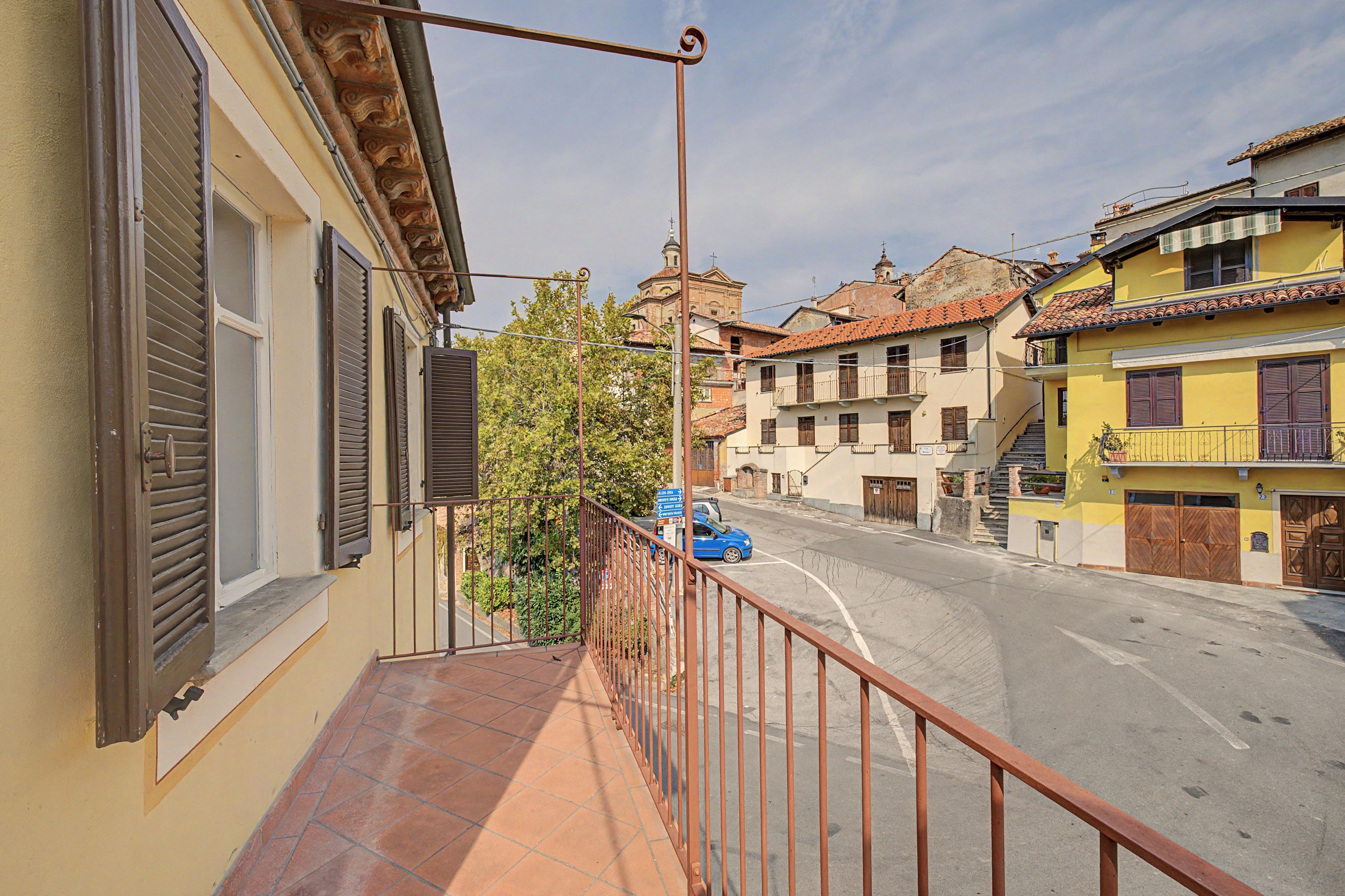 Un balcon doté d’une rambarde en métal donnant sur une rue bordée d’immeubles, avec une voiture bleue garée sur le bord de la route.