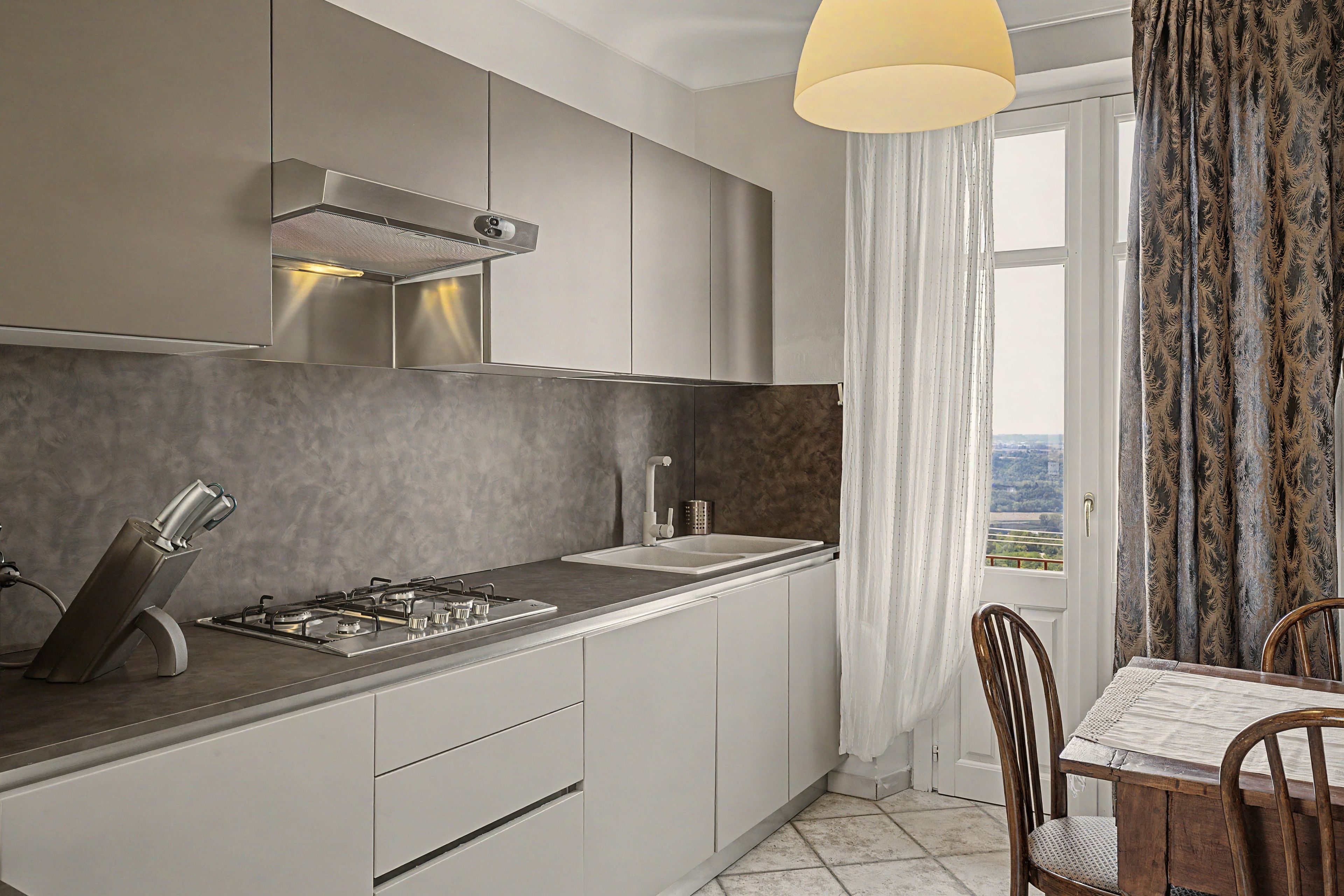 A modern kitchen with a gas stove, white cabinets, a stainless steel range hood, and a view through a window with curtains.