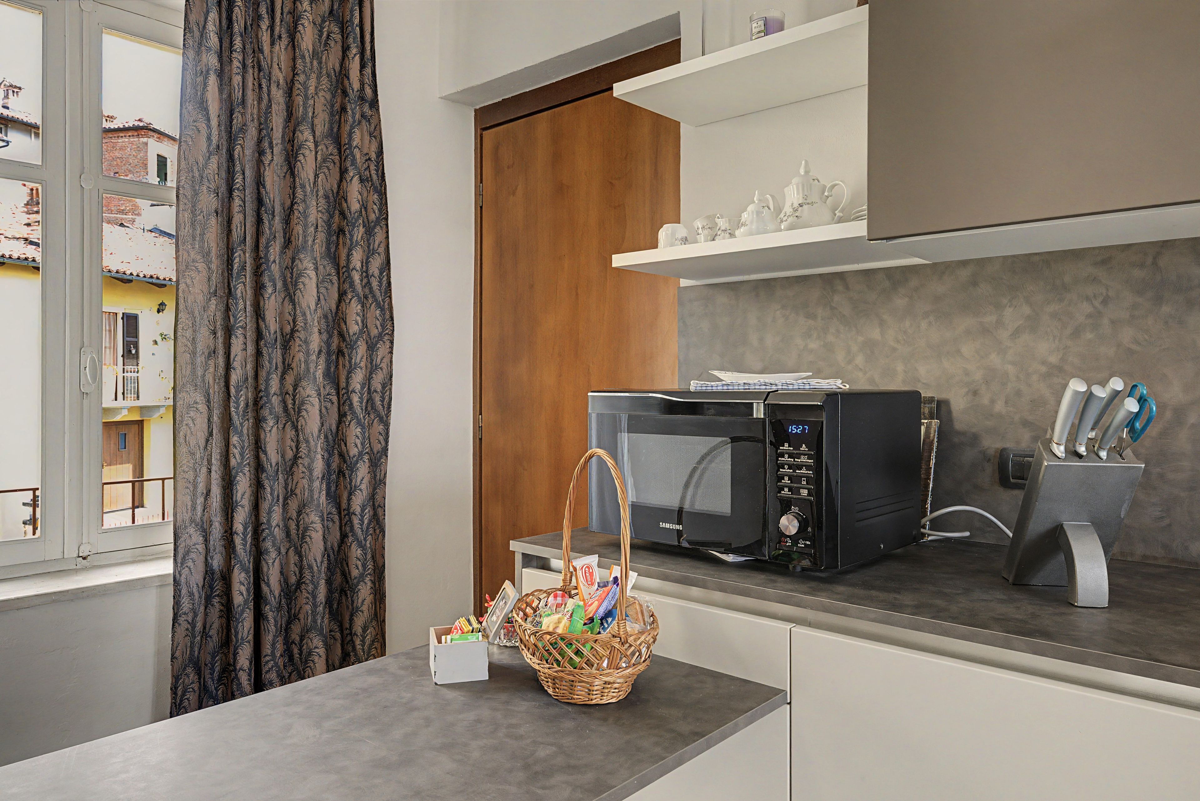 A modern kitchen with a microwave, a basket of groceries, and a window with curtains.