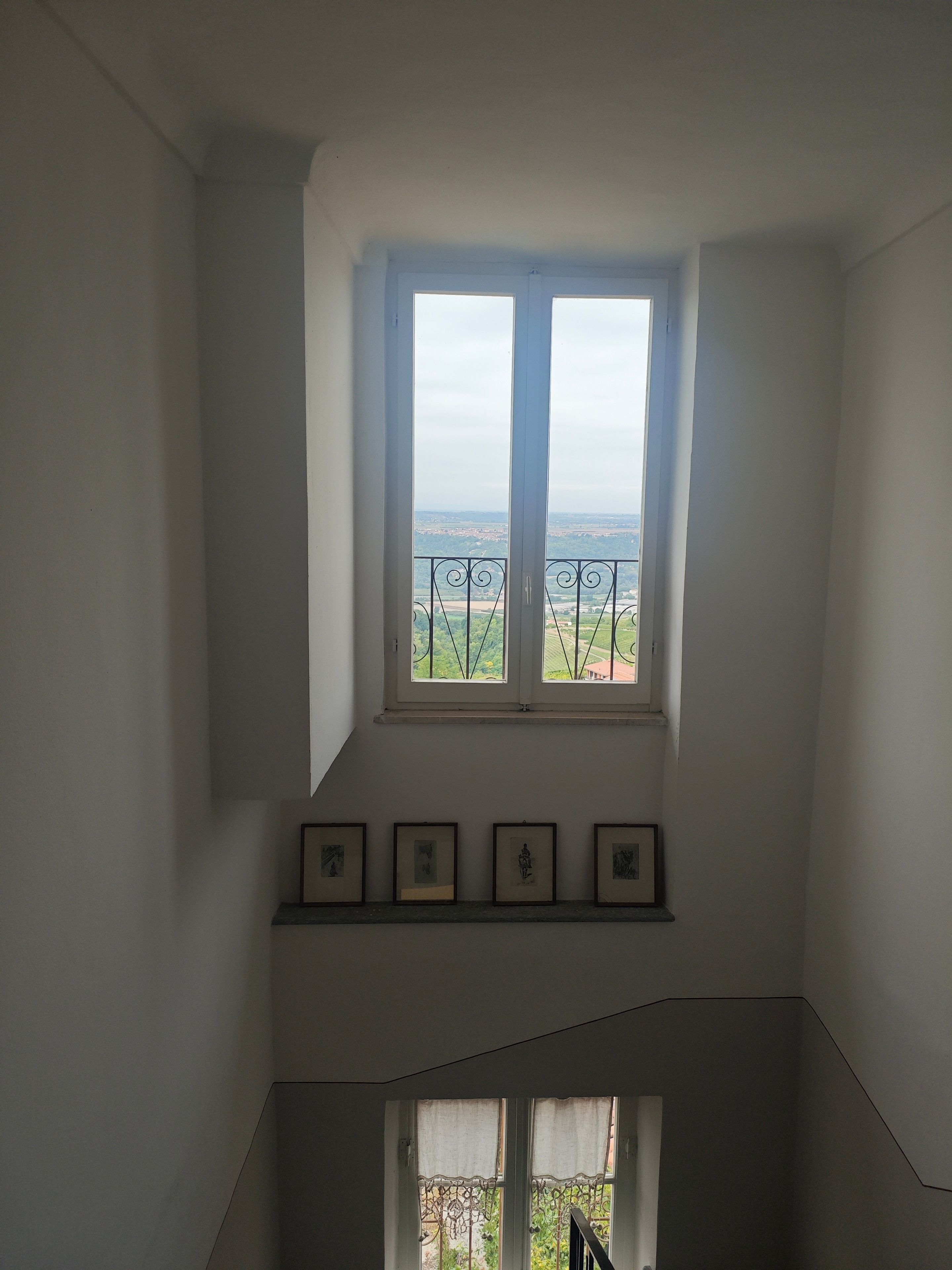 Ein Zimmer mit Fensterblick auf eine Landschaft, einer Treppe und mehreren gerahmten Bildern an der Wand.