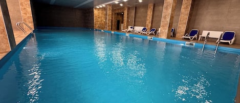 An indoor swimming pool with clear water, a tiled floor, and lounge chairs along the wall.