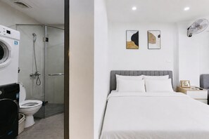 Quarto de hotel com cama, mesa de cabeceira, ventoinha na parede e área de chuveiro com porta de vidro.