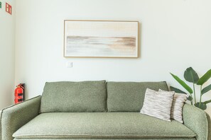 A green sofa with a fire extinguisher and a plant in a room with a white wall and a framed artwork. - Estrelicia Studio by Atlantic Holiday (Santa Cruz)