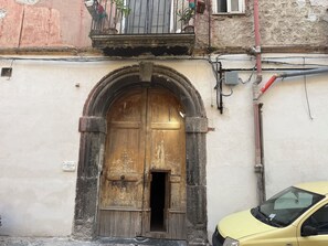 Una puerta de madera desgastada con un arquitrabe de metal, un balcón con macetas y un coche amarillo estacionado cerca.