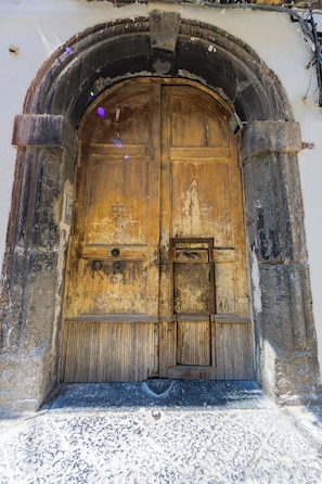 Una puerta de madera grande con un pequeño ventanal y un pestillo de metal, ubicada en una estructura de piedra en arco.