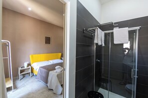 Habitación de hotel con cama, mesita de noche, zona de ducha y perchero.