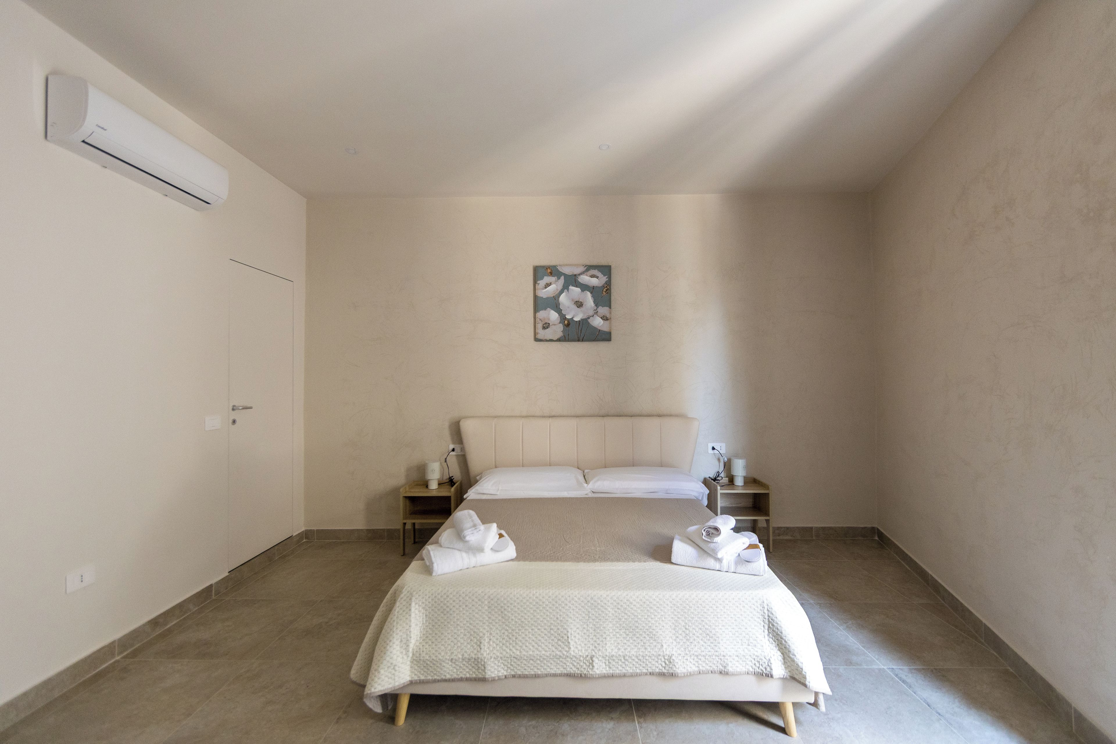 Ett minimalistiskt hotellrum med en säng, ett sängbord, en väggmonterad luftkonditioneringsanläggning och ett enda konstverk.