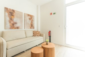 A modern living room with a sofa, two round wooden side tables, abstract wall art, a fire extinguisher mounted on the wall, and a door with an exit sign. - Orquídea Studio by Atlantic Holiday (Santa Cruz)