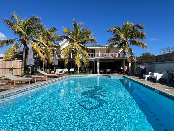 En swimmingpool med liggestole og parasoller, omgivet af palmer og en bygning med balkon.