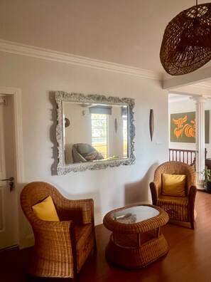 Ein Zimmer mit zwei Korbsesseln, einem kleinen runden Tisch und einem großen Spiegel mit aufwendiger Rahmung.