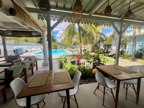 Un patio al aire libre con mesas de madera y sillas blancas, con vistas a una piscina y abundante follaje.