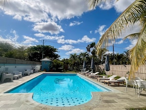 Et rektangulært swimmingpool med flisebelagt solarium, omgivet af liggestole og parasoller, under en klar himmel.