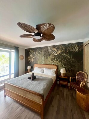 A bedroom with a bed, a ceiling fan, a wall with a tropical leaf mural, a window, and a chair. - Lakaz Poz 5 - Chambre double - 200m de la mer (Albion)