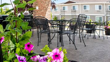 An outdoor patio with metal chairs and tables, surrounded by potted plants and a brick pillar.