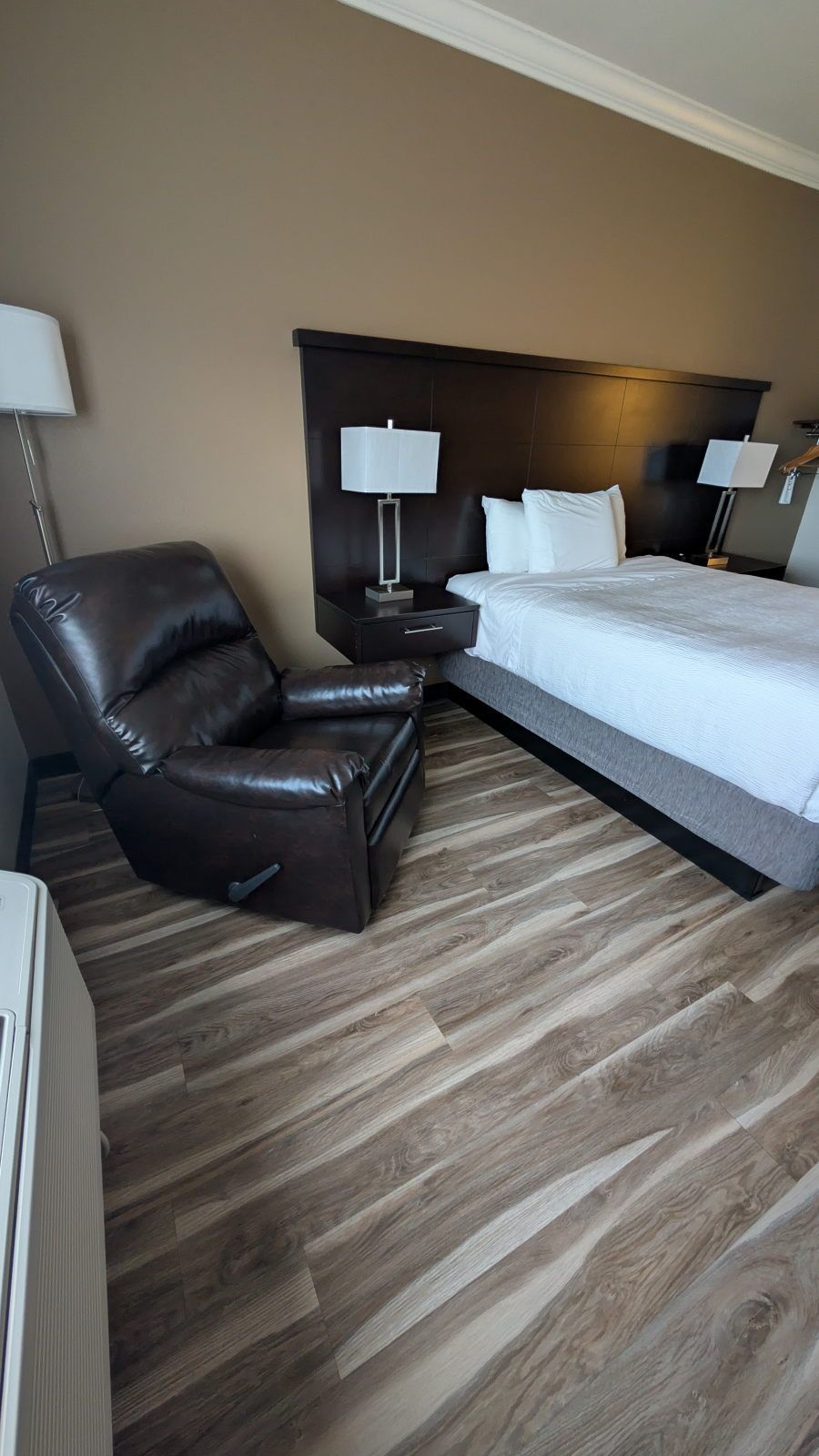 Una habitación de hotel con una cama, una butaca reclinable, una mesita de noche con lámpara y piso de madera.