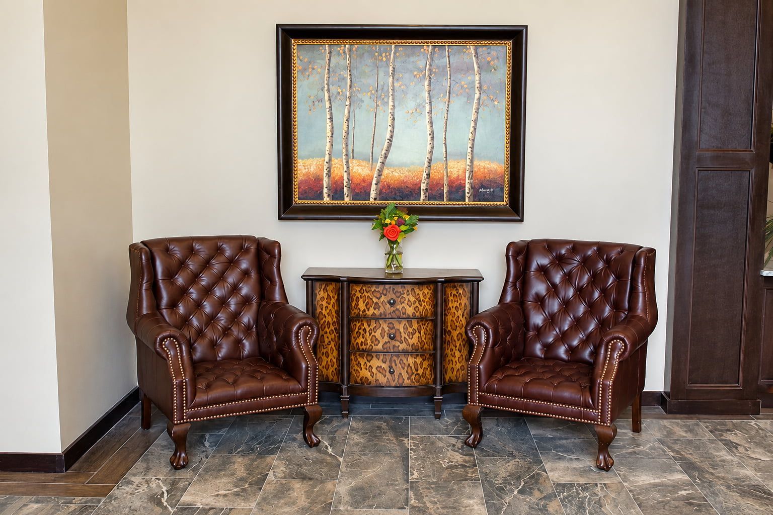 Two brown leather armchairs with tufted backs, a small table with a vase of flowers, and a framed painting on the wall.