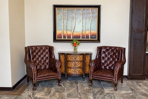 Two brown leather armchairs with tufted backs, a small table with a vase of flowers, and a framed painting of trees on the wall. - ADA Queen Room w/ Kitchen & Free Breakfast (Moses Lake)