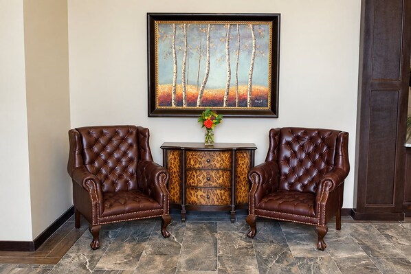 Two brown leather armchairs with tufted backs, a small table with a vase of flowers, and a framed painting of trees on the wall.
