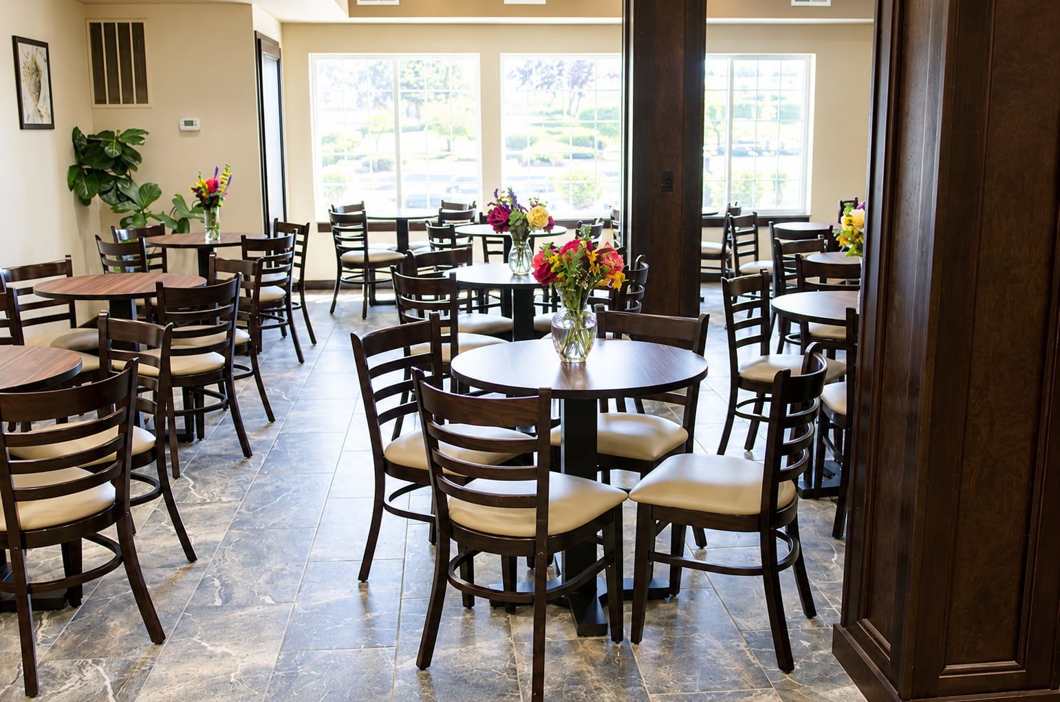 A spacious dining area with multiple round tables and chairs, large windows, and potted plants.