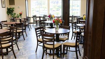 A spacious dining area with multiple round tables and chairs, large windows, and potted plants.