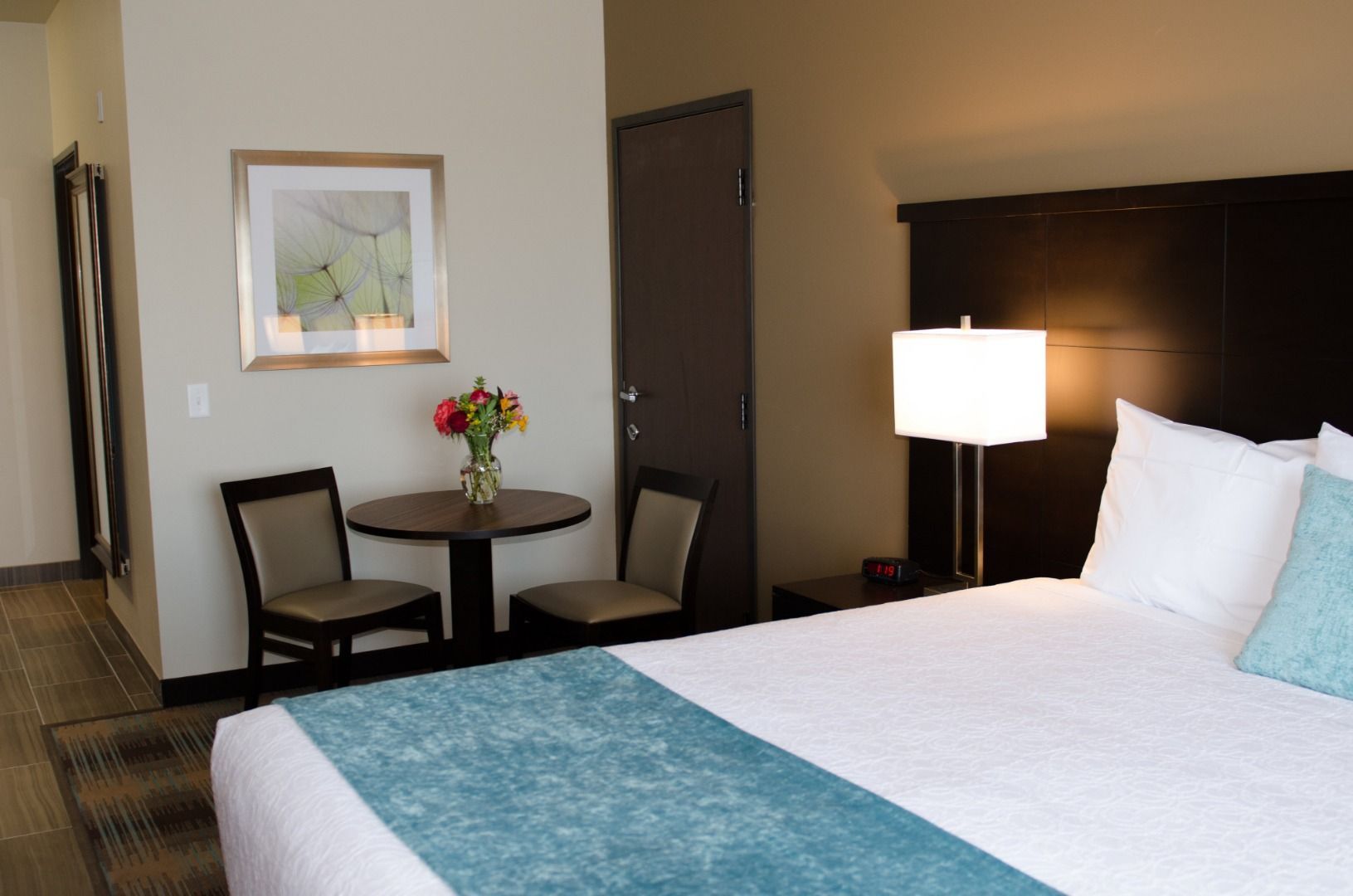 A hotel room with a bed, a bedside lamp, a small table with chairs, a vase with flowers, and a framed picture on the wall.