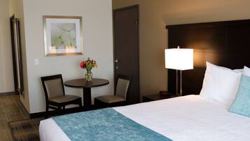 A hotel room with a bed, a bedside lamp, a small table with chairs, a vase with flowers, and a framed picture on the wall.