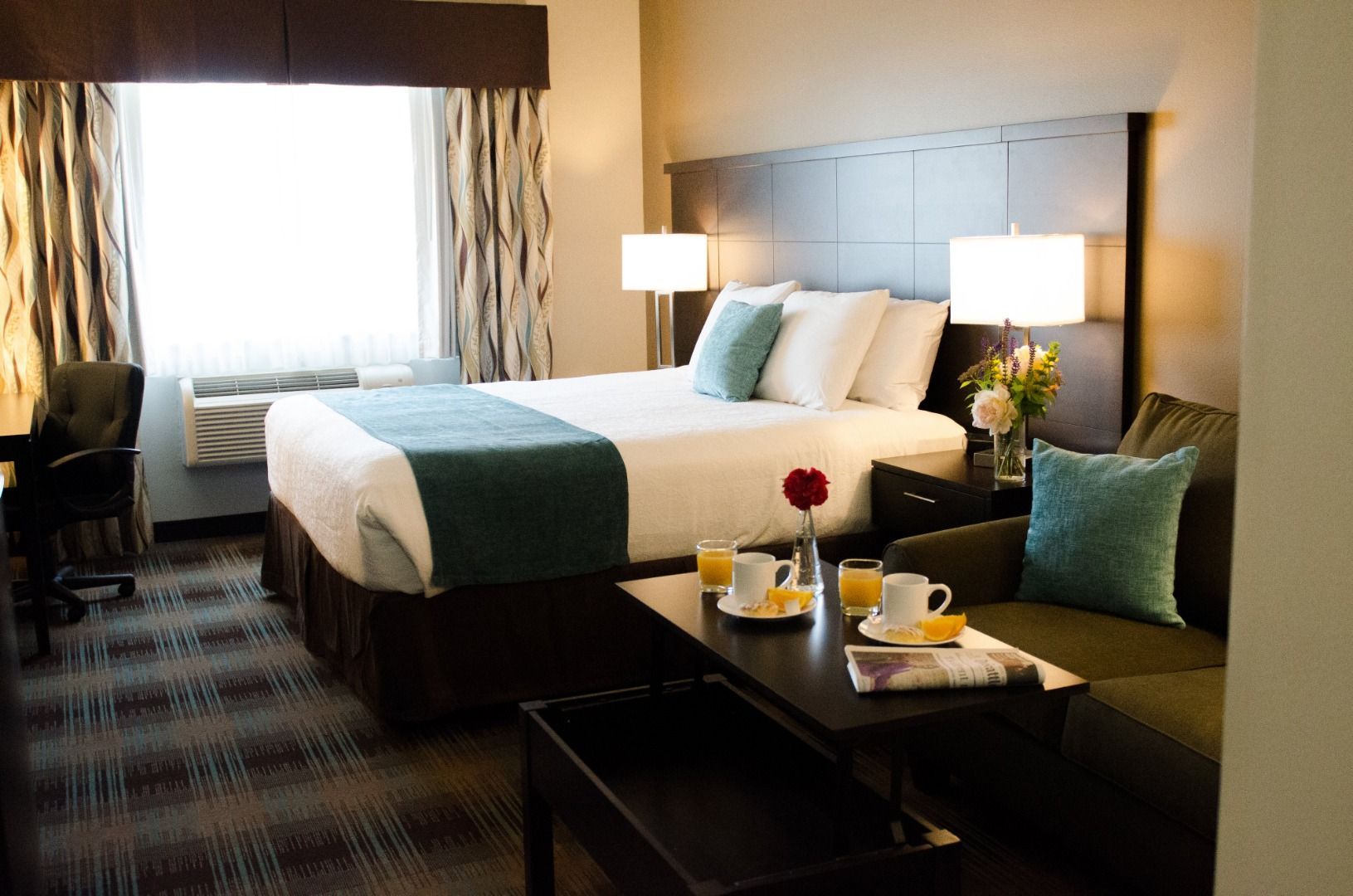 A hotel room with a bed, two bedside lamps, a coffee table with breakfast items, a chair, and a desk.