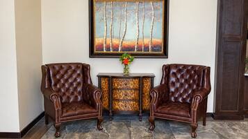 Two brown leather armchairs with tufted backs, a small table with a vase of flowers, and a framed painting of trees on the wall.