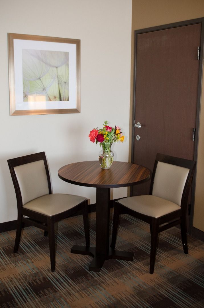 Een hotelkamer met een ronde houten tafel, twee stoelen, een vaas met bloemen, een lijst met een afbeelding aan de muur en een gesloten deur.