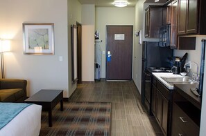 Habitación de hotel compacta con cama, mesita de noche y un pequeño escritorio. Un pasillo conduce a una kitchenette con refrigerador, microondas y lavamanos.