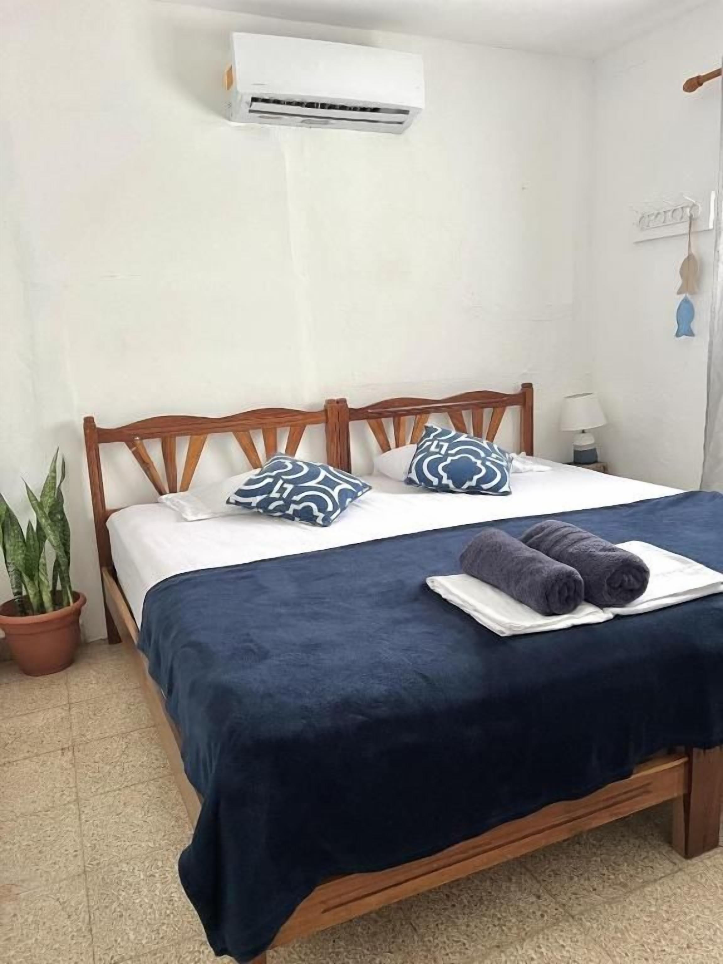 Une chambre à coucher avec un lit en bois, une couvre-lit bleu foncé, deux oreillers, une serviette pliée, un appareil de climatisation, une lampe de chevet et une plante en pot.