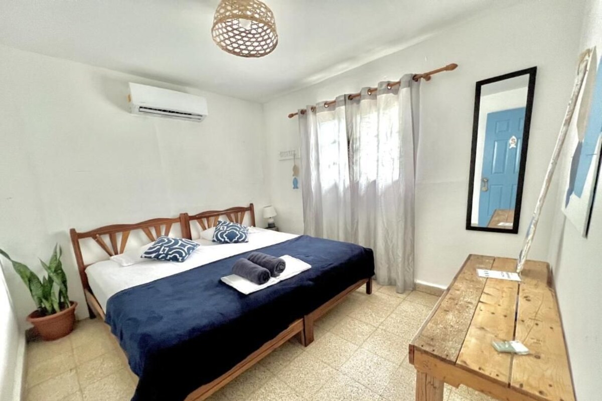 A bedroom with a bed, a wooden table, a potted plant, and a window with curtains.