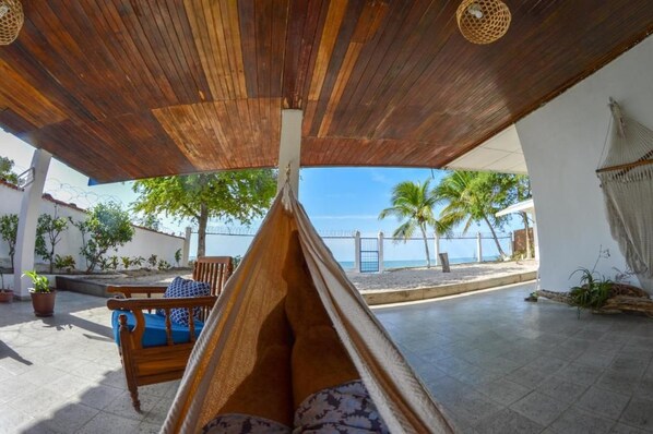 A wooden ceiling with a hammock, a wooden chair with a cushion, and a view of the beach and palm trees.