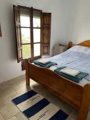 A single bed with a wooden frame, a window with open shutters, and a blue-striped rug on a tiled floor. - Casa Blanca | Lecrin Valley, Granada (Acequias)