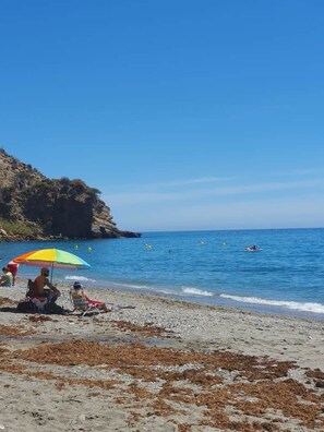 Ein Strand mit Menschen unter Sonnenschirmen, eine felsige Klippe und ruhiges Meer.