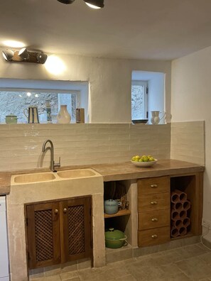 A kitchen with a sink, wooden cabinets, a window, and a bowl of fruit on the counter. - Casa Blanca | Lecrin Valley, Granada (Acequias)